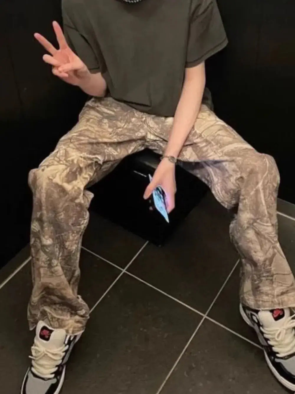 Man sitting on a black stool wearing Twig Camouflage Distressed Jeans and sneakers, stylish casual outfit.